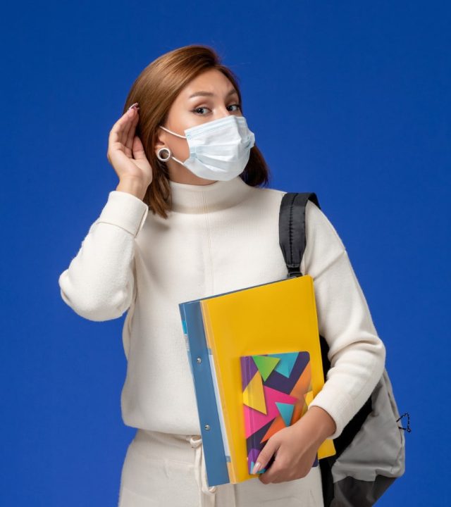 front-view-young-female-student-white-jersey-wearing-mask-with-bag-copybooks-trying-hear-blue-wall-min